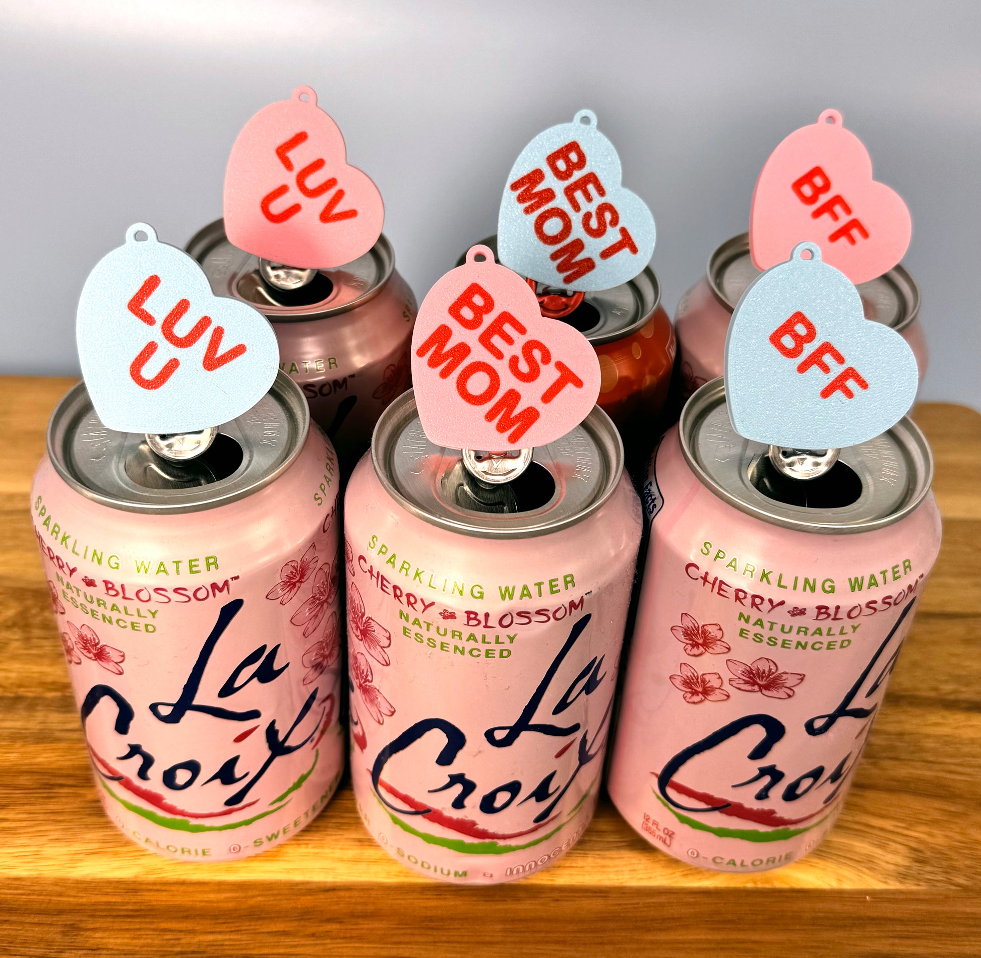 Pink La Croix cans with pink and blue Candy Heart Tab Buddies with vaarious phrases on a wooden surface.