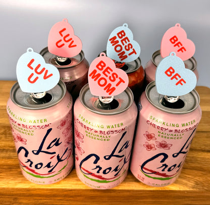 Pink La Croix cans with pink and blue Candy Heart Tab Buddies with vaarious phrases on a wooden surface.