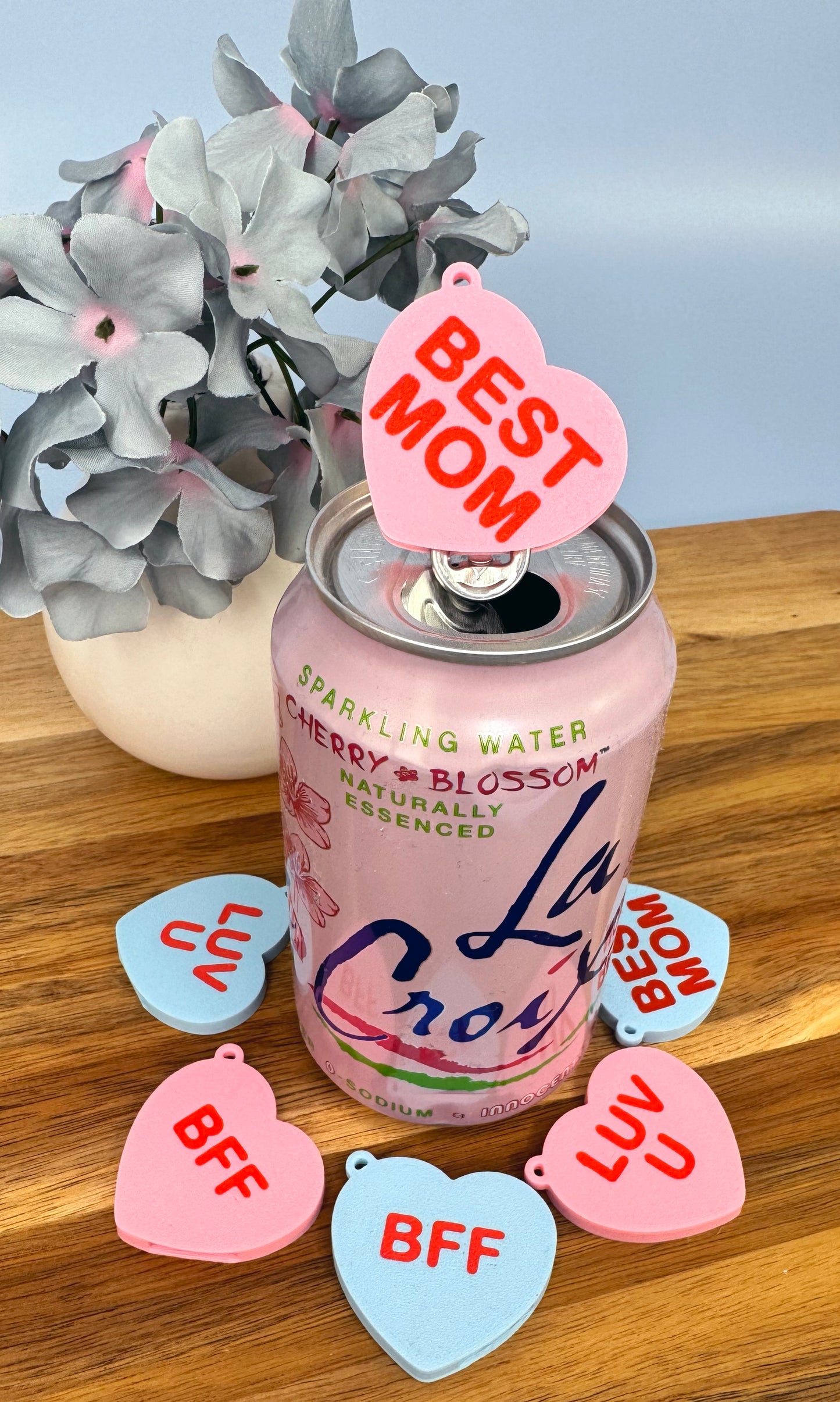 Pink can of La Croix sparkling water with 'BEST MOM' and heart-shaped Tab Buddies on a wooden surface with flowers in the background.