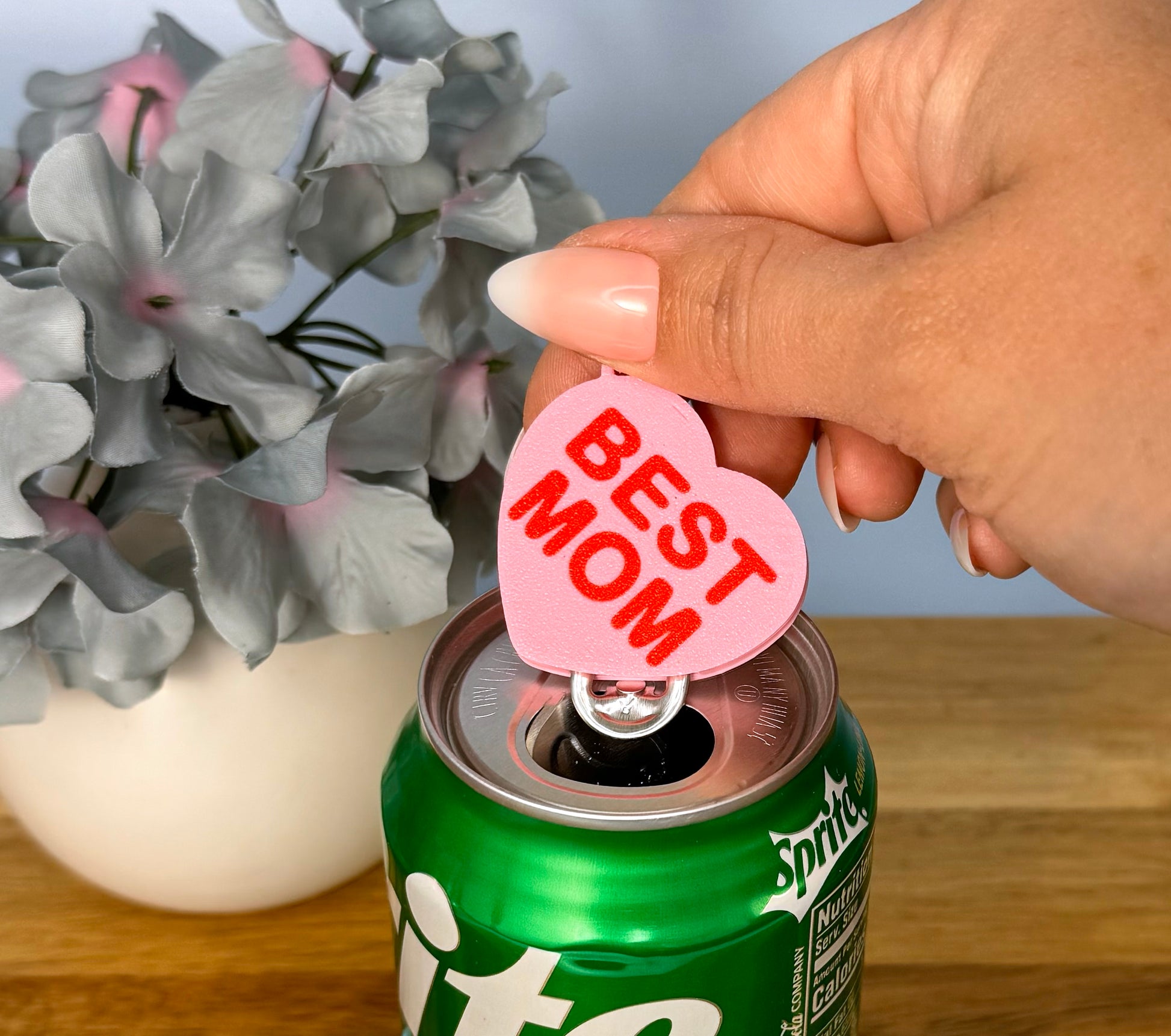Hand using a 'Best Mom' Tab Buddy on a Sprite can with flowers in the background