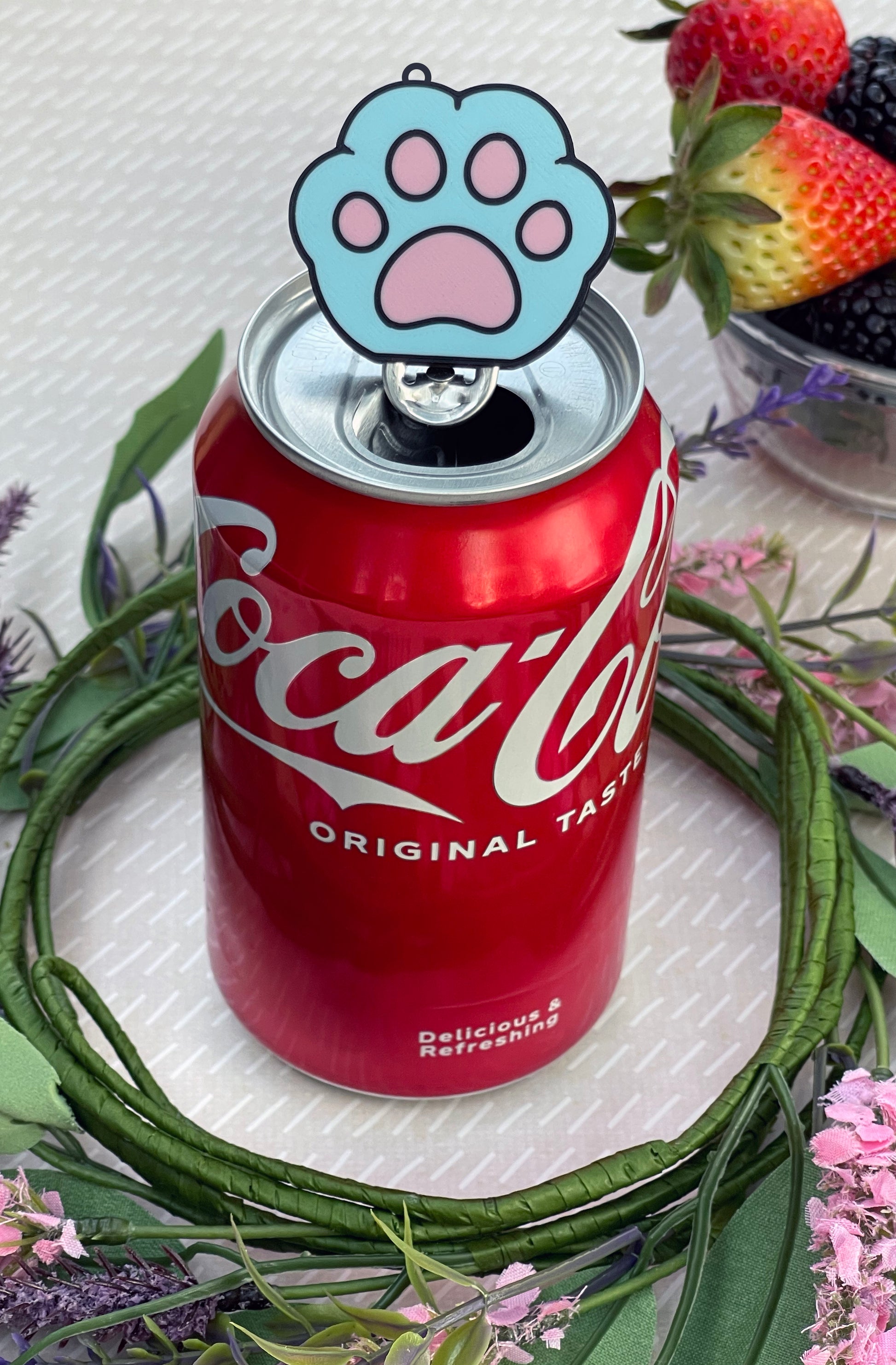 Red Coca-Cola can with a blue paw print Tab Buddy resting on top, surrounded by flowers and berries.