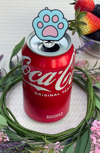Red Coca-Cola can with a blue paw print Tab Buddy resting on top, surrounded by flowers and berries.