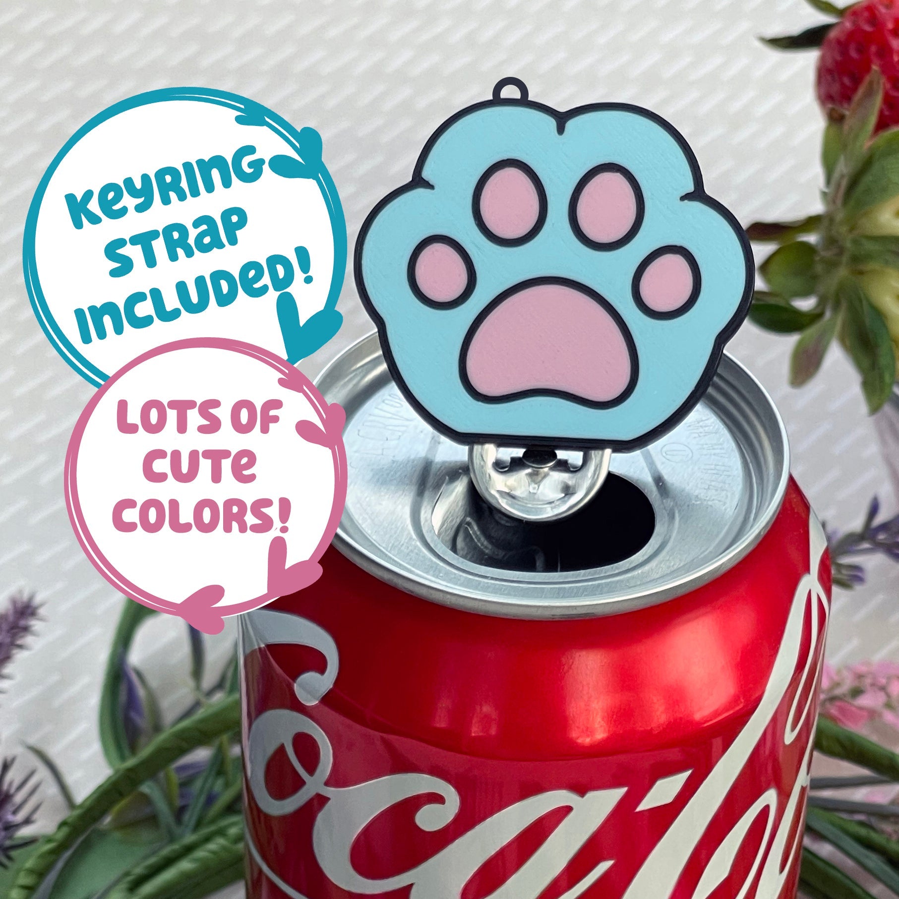 Coca-Cola can with a blue and pink paw print Tab Buddy keychain on top, surrounded by plants.