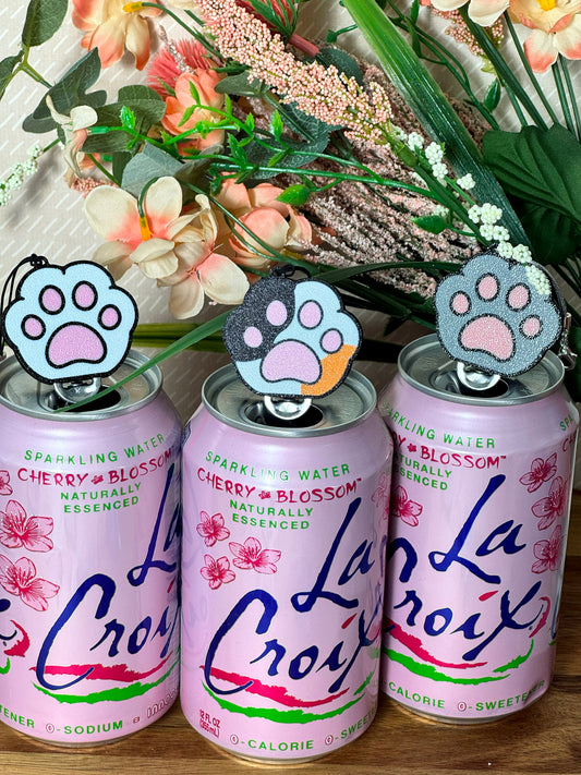 Three La Croix sparkling water cans with colorful Kitty Paw Tab Buddies on a wooden surface with flowers in the background.