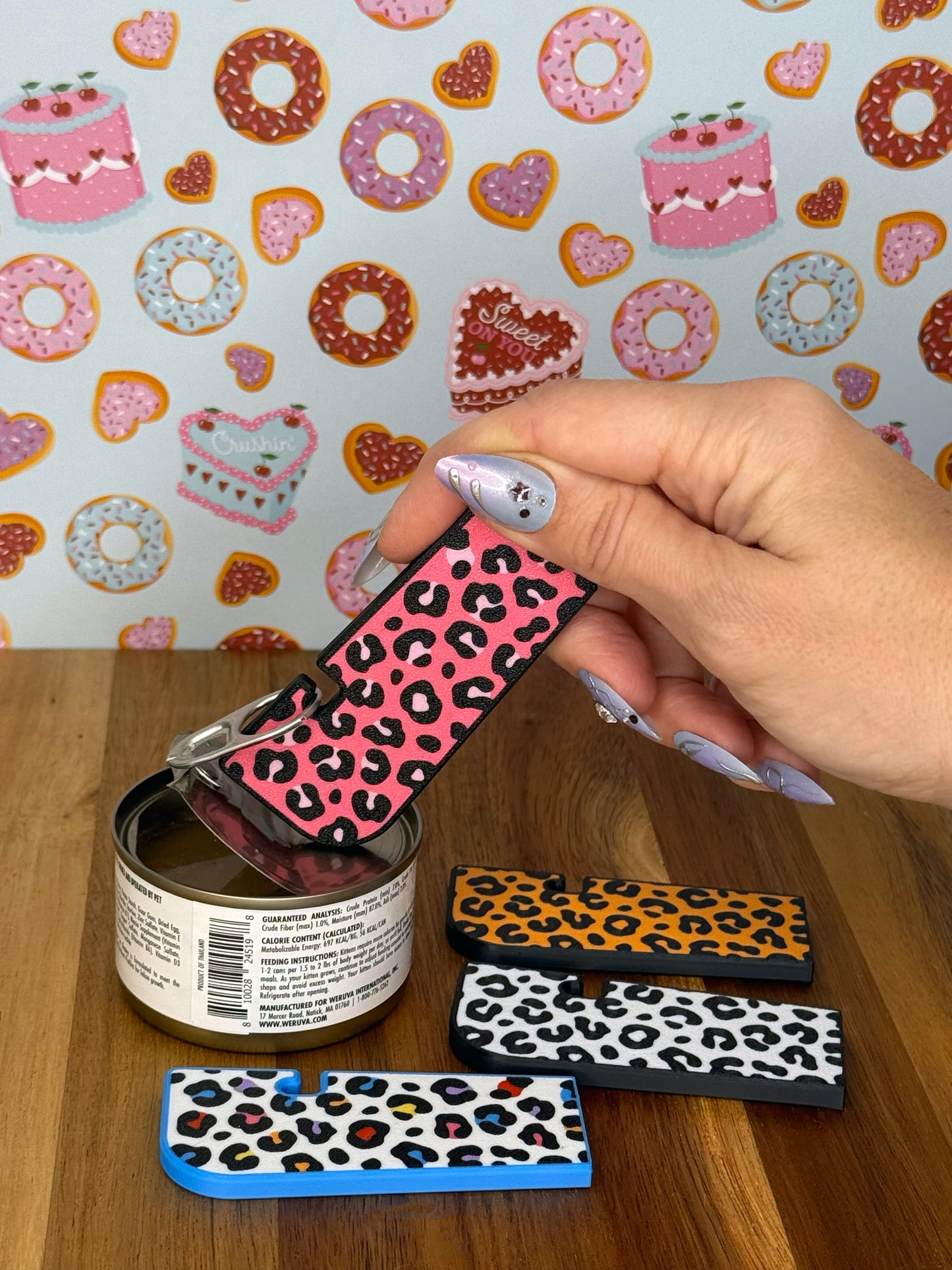 Hand holding a Tab Buddy XL with a pink leopard print design against a colorful background. Three more color options for the leopard print design rest on a wooden surface.