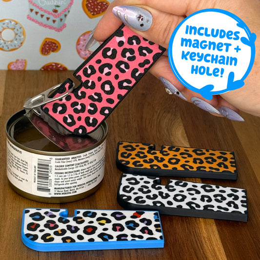 Hand holding a Tab Buddy XL with a pink leopard print design against a colorful background. Three more color options for the leopard print design rest on a wooden surface. Text box reads, "Includes magnet + keychain hole!"