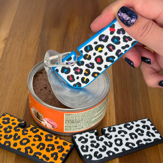 A rainbow leopard print Tab Buddy XL is used to open a can of cat food with additional leopard print Tab Buddies on a wooden surface.