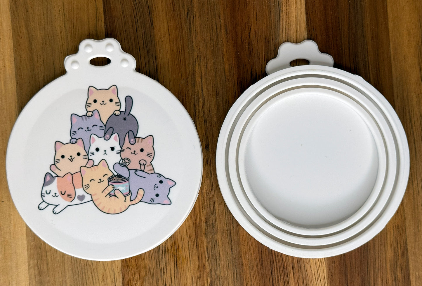 The front and back of a reusable silicone can lid with a cute cat design are shown resting atop a wooden surface.