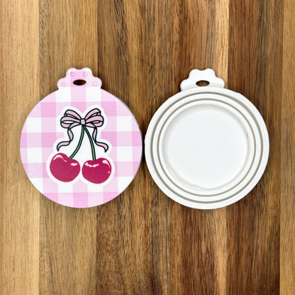 The front and back of a reusable silicone can lid with a cute pink gingham and cherries design are shown resting atop a wooden surface.