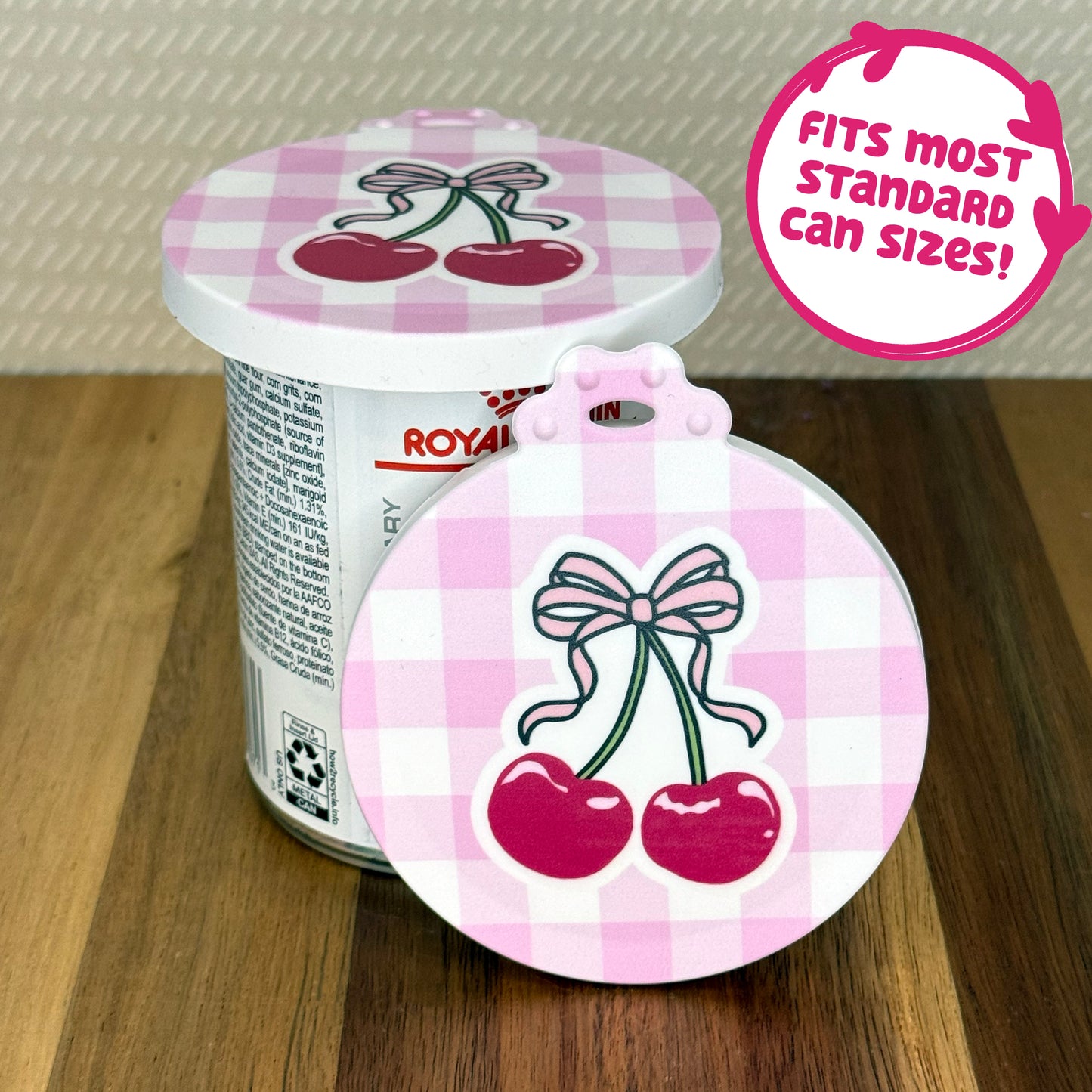 A cute reusable silicone can lid featuring a design of a two cherries with a bow on top and a pink gingham background is shown fitted atop a can. Text reads "Fits most standard can sizes!"