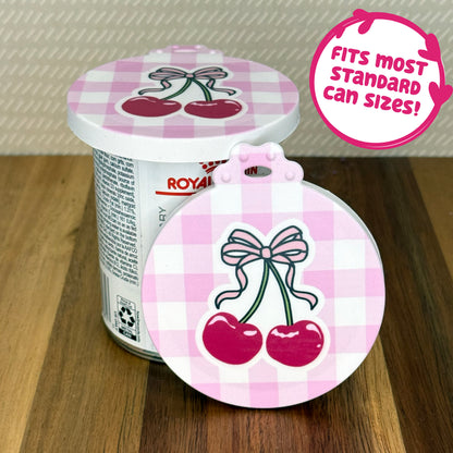 A cute reusable silicone can lid featuring a design of a two cherries with a bow on top and a pink gingham background is shown fitted atop a can. Text reads "Fits most standard can sizes!"