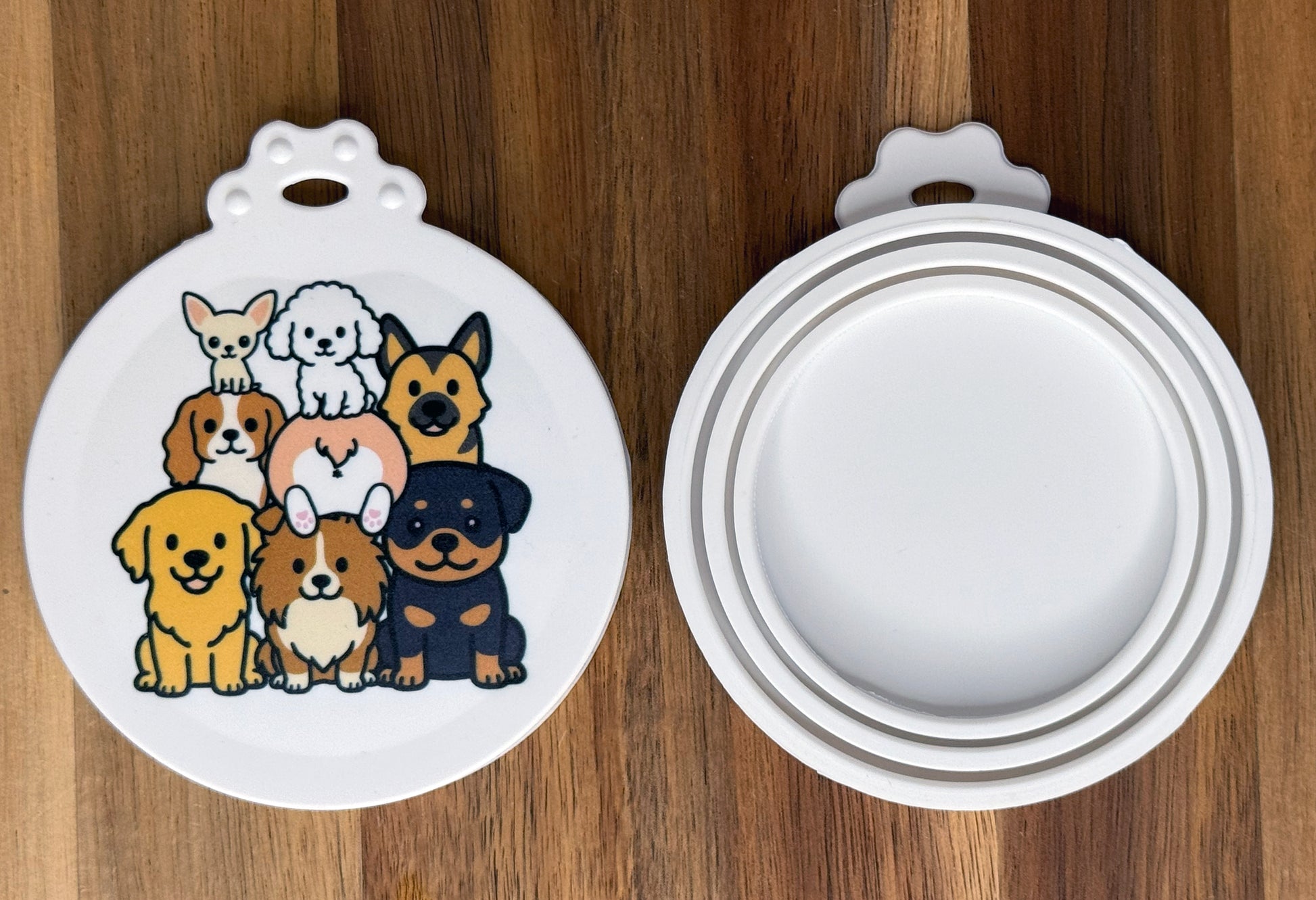 The front and back of a reusable silicone can lid with a cute dog design are shown resting atop a wooden surface.