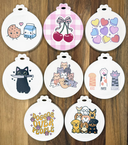 A group of 8 Tab Buddies reusable can lids available in Spring 2026 are shown on a wooden surface. Designs include themes of food, cats and dogs.