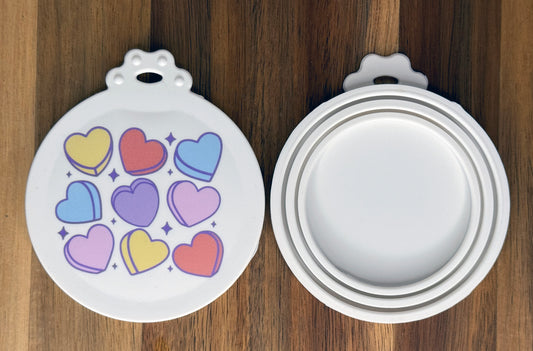 The front and back of a reusable silicone can lid with a cute Candy Hearts design are shown resting atop a wooden surface.