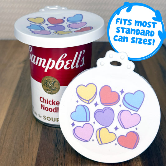 A can lid featuring cute, colorful candy hearts rests in front of a can of soup with another lid covering the top. Descriptive text reads, "fits most standard can sizes!"