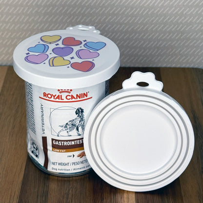 A can of dog food is sealed with a resusable silicone can cover with hearts design. An additional lid is shown facing outward with multiple circular rings that stretch to seal various sized cans.