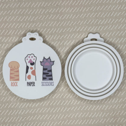 The front and back of a reusable silicone can lid with a cute cat paw design reading "rock paper scissors" are shown resting atop a beige background.