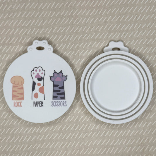 The front and back of a reusable silicone can lid with a cute cat paw design reading "rock paper scissors" are shown resting atop a beige background.