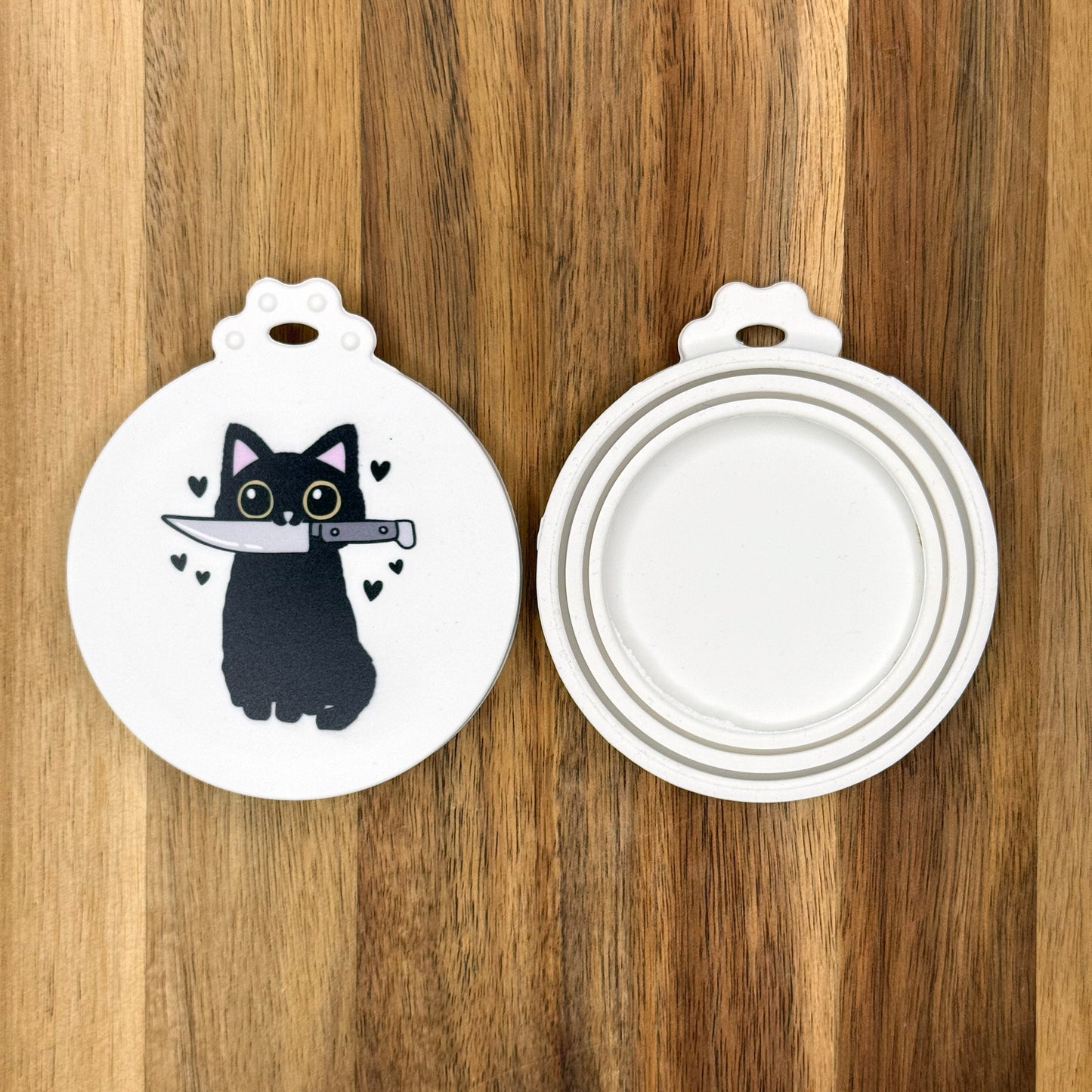 The front and back of a reusable silicone can lid with a cute design of a cat holdionig a knife in its mouth are shown resting atop a wooden surface.