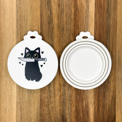 The front and back of a reusable silicone can lid with a cute design of a cat holdionig a knife in its mouth are shown resting atop a wooden surface.