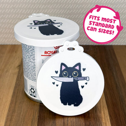 A cute reusable silicone can lid featuring a design of a cartoon-style black cat holding a knife in its mouth with bleack hearts surrounding it is shown fitted atop a can. Text reads "Fits most standard can sizes!"