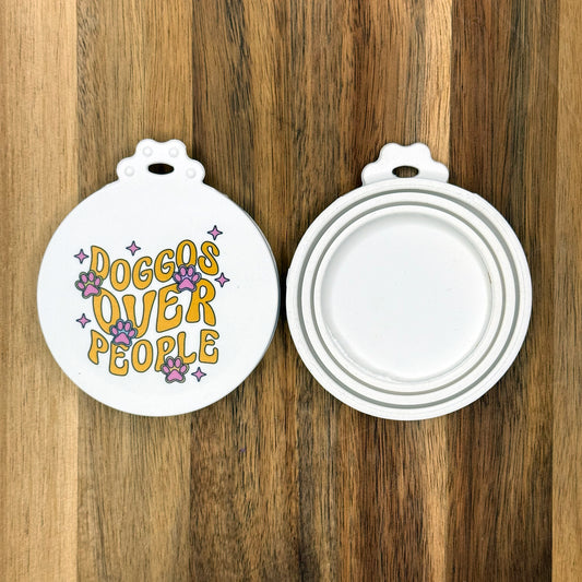 The front and back of a reusable silicone can lid with a groovy-styled design with paws, twinkles and text reading "Doggos over people" are shown resting atop a wooden surface.