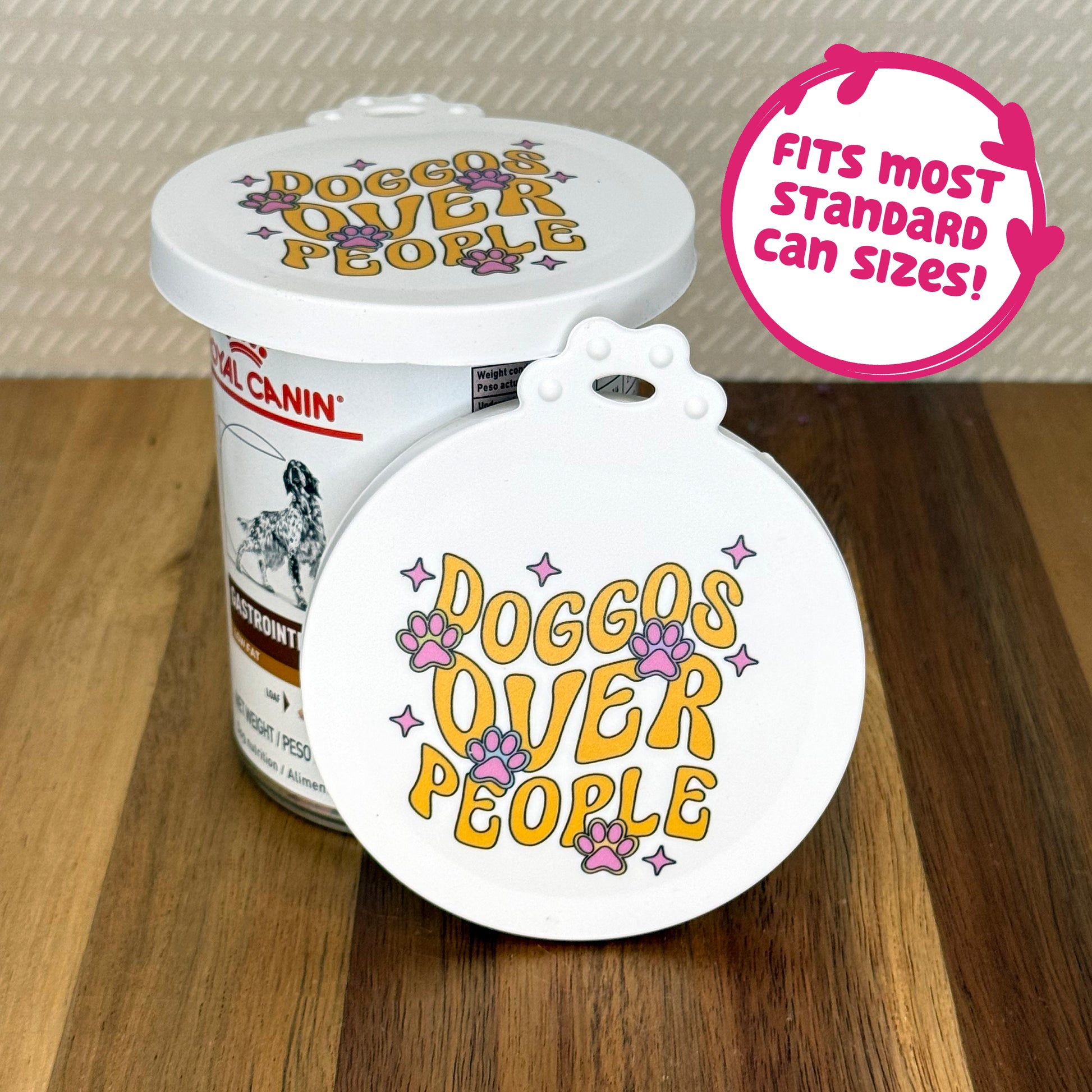 A cute reusable silicone can lid featuring a design of groovy-styled text reading "doggos over people" with paw prints and twinkles is shown fitted atop a can. Text reads "Fits most standard can sizes!"