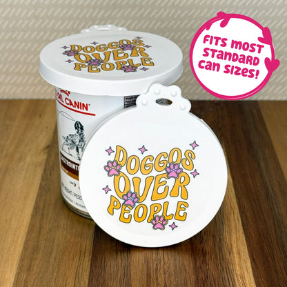 A cute reusable silicone can lid featuring a design of groovy-styled text reading "doggos over people" with paw prints and twinkles is shown fitted atop a can. Text reads "Fits most standard can sizes!"