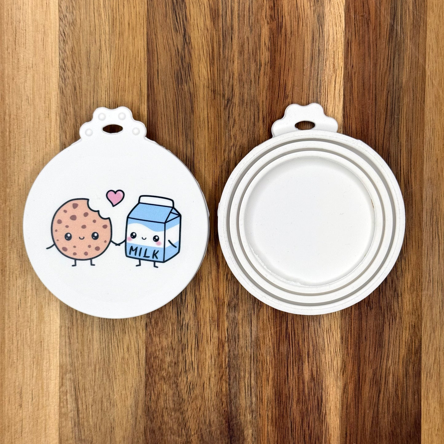 The front and back of a reusable silicone can lid with a cute milk and cookie design are shown resting atop a wooden surface.