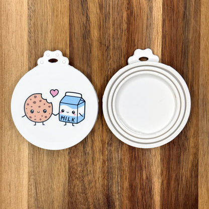 The front and back of a reusable silicone can lid with a cute milk and cookie design are shown resting atop a wooden surface.