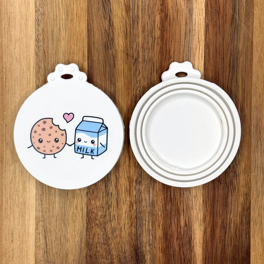 The front and back of a reusable silicone can lid with a cute milk and cookie design are shown resting atop a wooden surface.