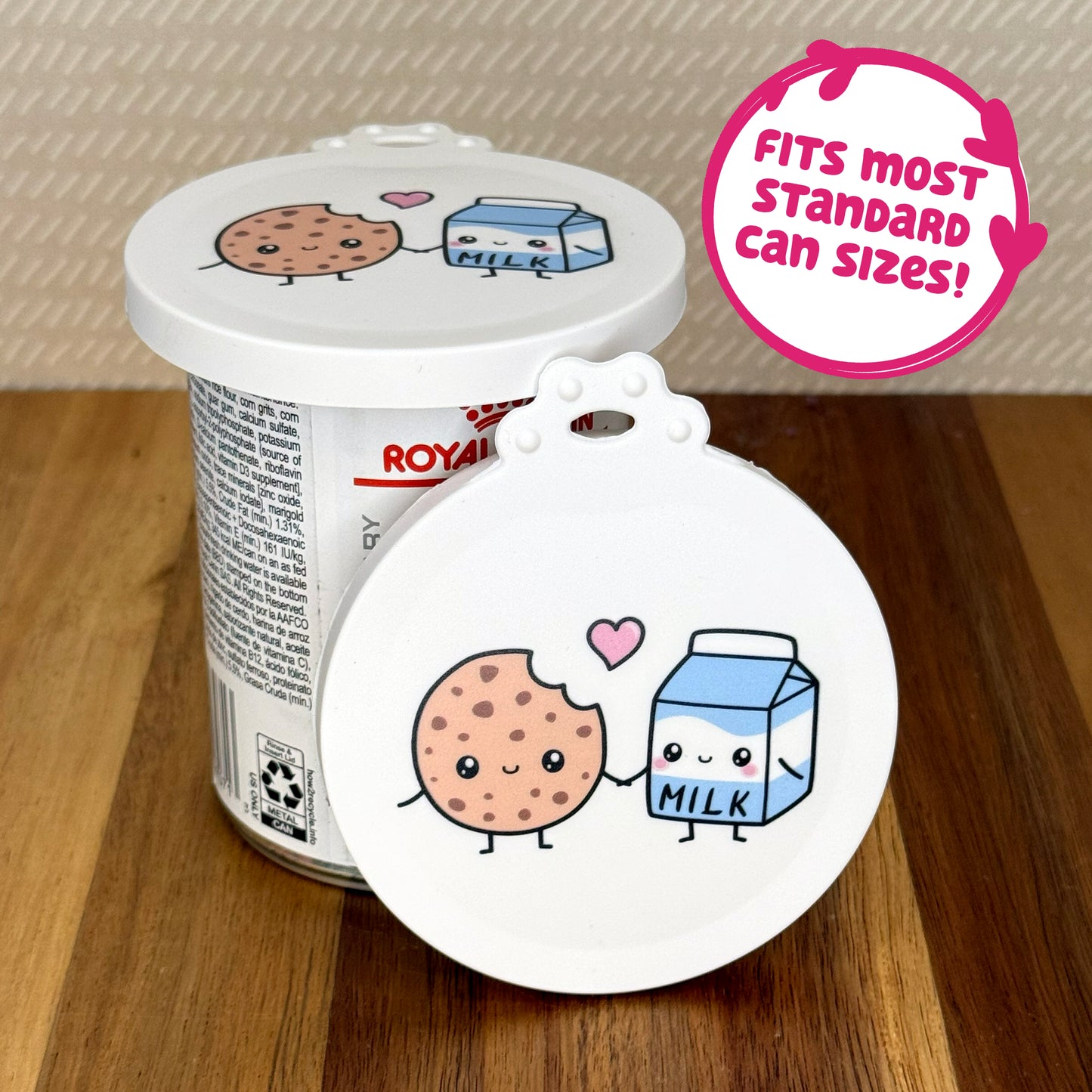A cute reusable silicone can lid featuring a design of a cartoon-style chocolate chip cooke and carton of milk holding hands is shown fitted atop a can. Text reads "Fits most standard can sizes!"