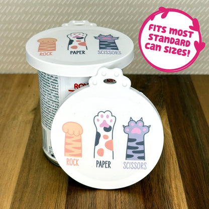 A cute reusable silicone can lid featuring a design of 3 cartoon-style cat paws with text underneath reading "rock, paper, scissors" is shown fitted atop a can. A text bubble reads "Fits most standard can sizes!"