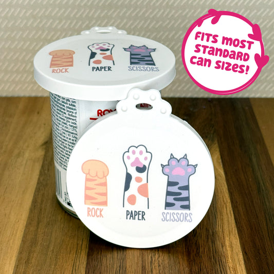 A cute reusable silicone can lid featuring a design of 3 cartoon-style cat paws with text underneath reading "rock, paper, scissors" is shown fitted atop a can. A text bubble reads "Fits most standard can sizes!"