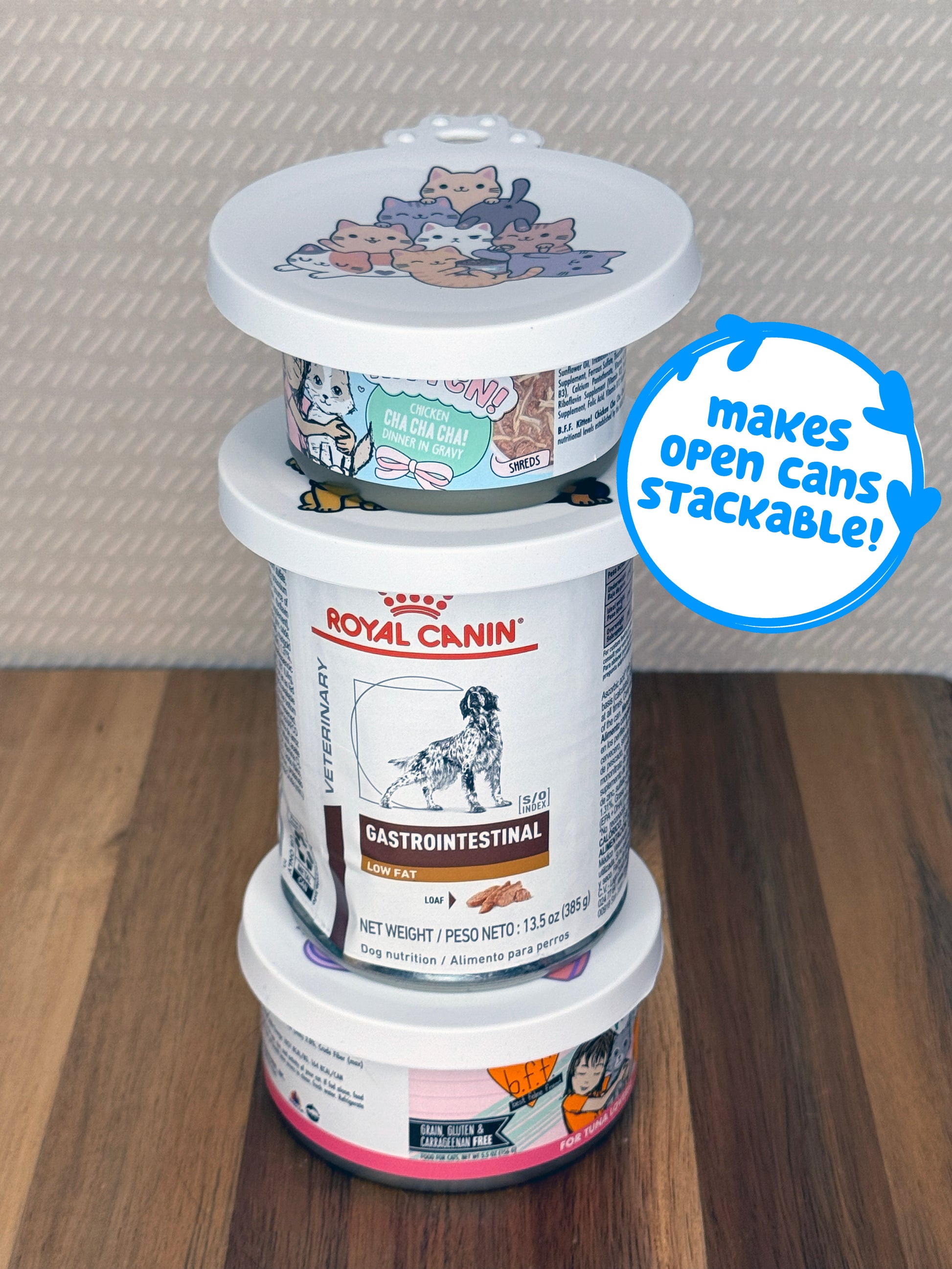 Stack of three pet food cans with a lid featuring cartoon cats, on a wooden surface. Descriptive text reads, "makes open cans stackable!"
