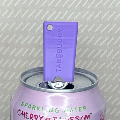 A purple Tab Buddy Mini soda can opener rests atop the tab of an open can of sparkling water with a beige background.