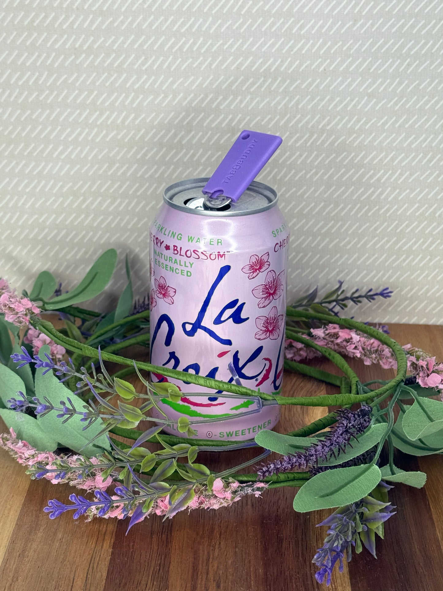 A purple Tab Buddy Mini soda can opener rests atop the tab of an open can of sparkling water with a beige background and floral accents.