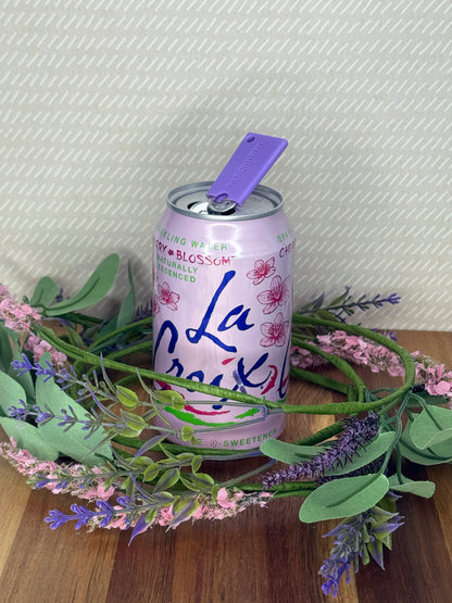 A purple Tab Buddy Mini soda can opener rests atop the tab of an open can of sparkling water with a beige background and floral accents.