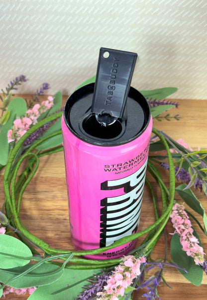 Pink energy drink can with black Tab Buddy Mini on a wooden surface surrounded by greenery and flowers