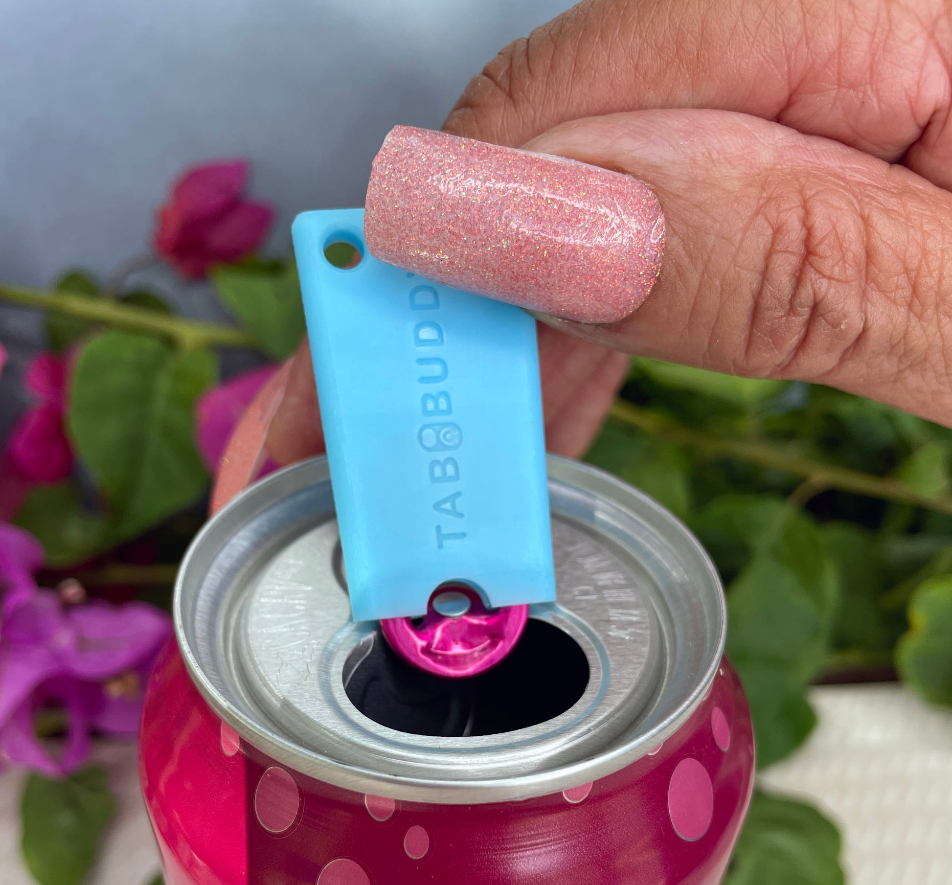 Hand using a blue can Tab Buddy Mini on a pink can with a blurred floral background