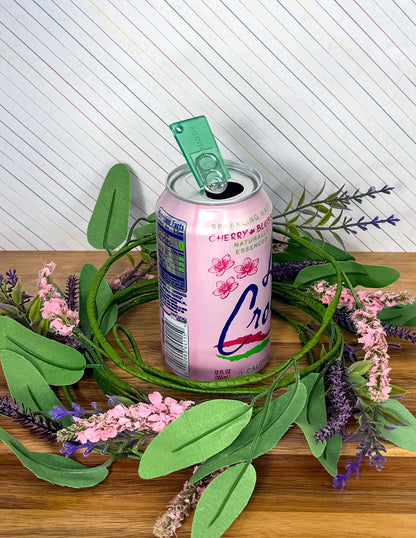 Pink can with an emerald green translucent Tab Buddy Mini on a wooden surface surrounded by green leaves and pink flowers.