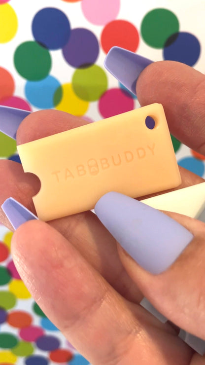 Hand holding a small peach-colored rectangular Tab Buddy Mini against a colorful polka dot background.