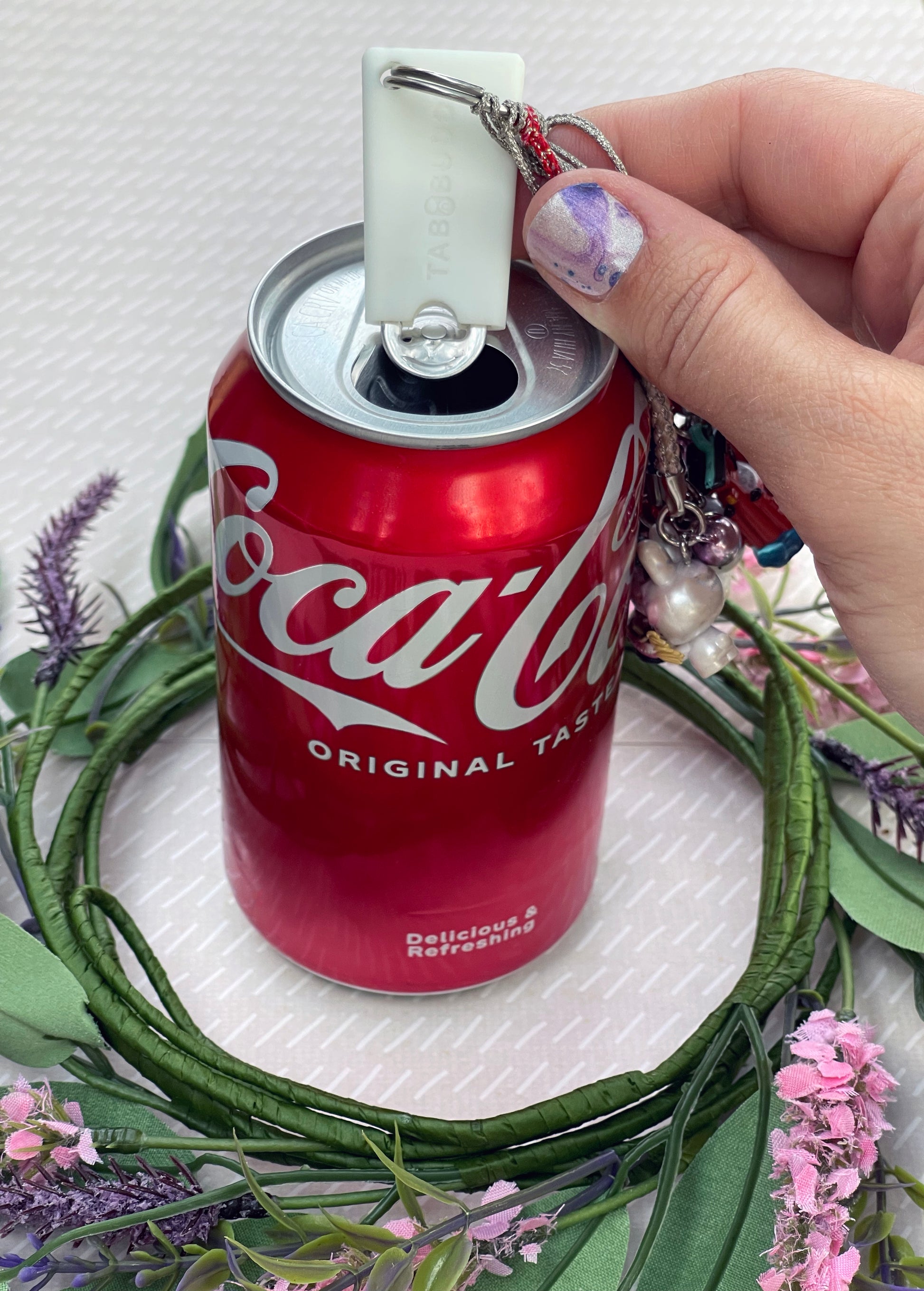 A white Tab Buddy Mini on a keychain sits atop a red Coca-Cola can surrounded by greenery and flowers.