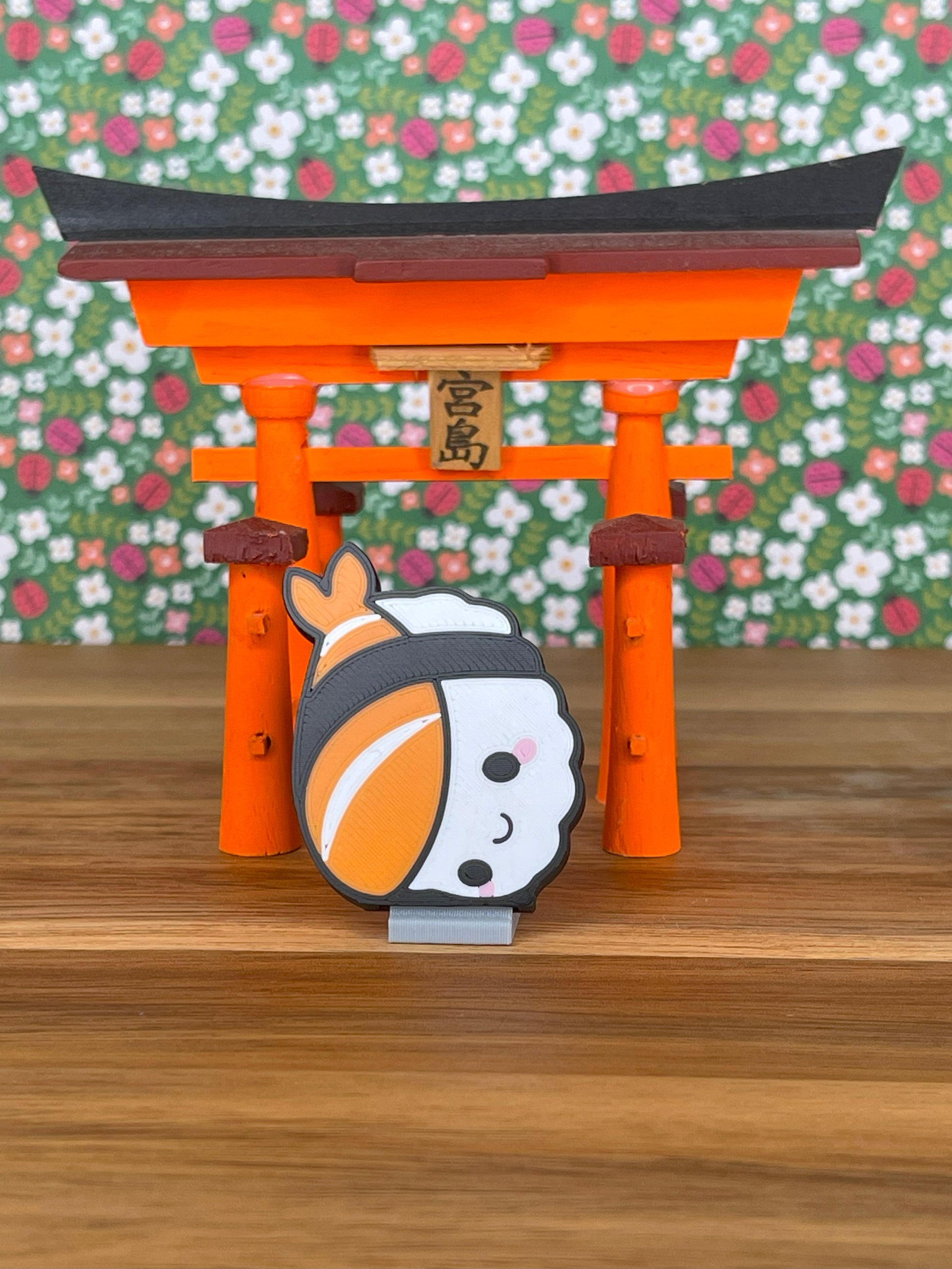Cute nigiri-shaped Tab Buddy stands in front of an orange torii gate on a wooden surface with floral wallpaper.