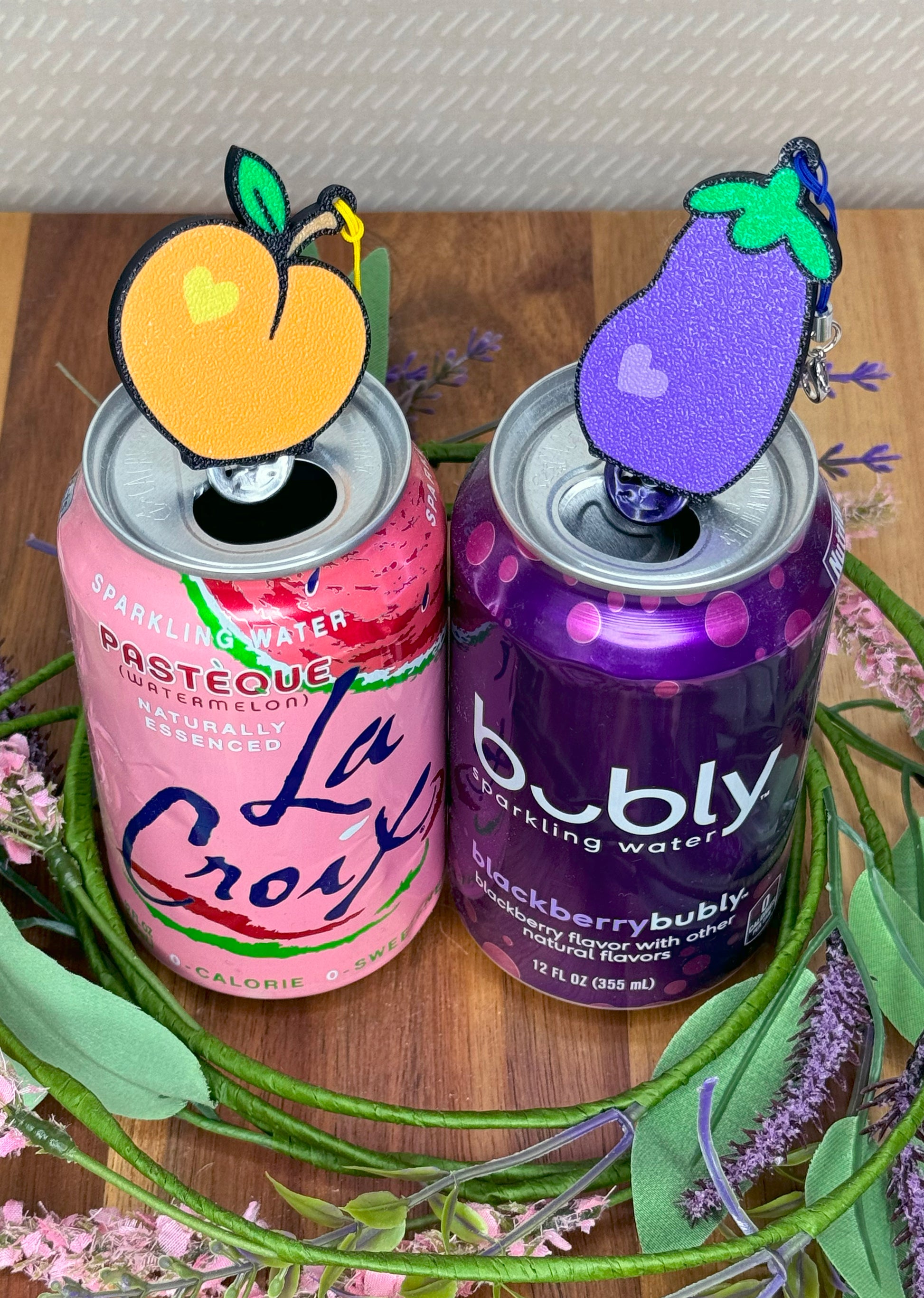 Two cans of sparkling water with decorative Tab Buddies on top of their tabs on a wooden surface.