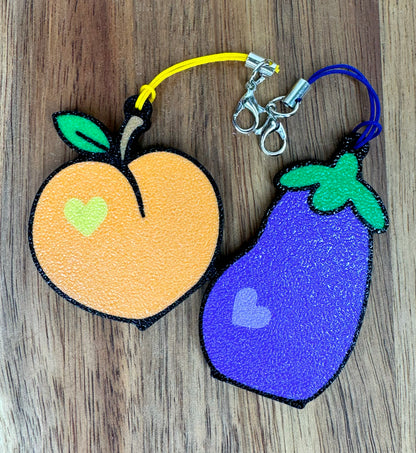 Two Tab Buddy keychains shaped like an peach and an eggplant on a wooden surface