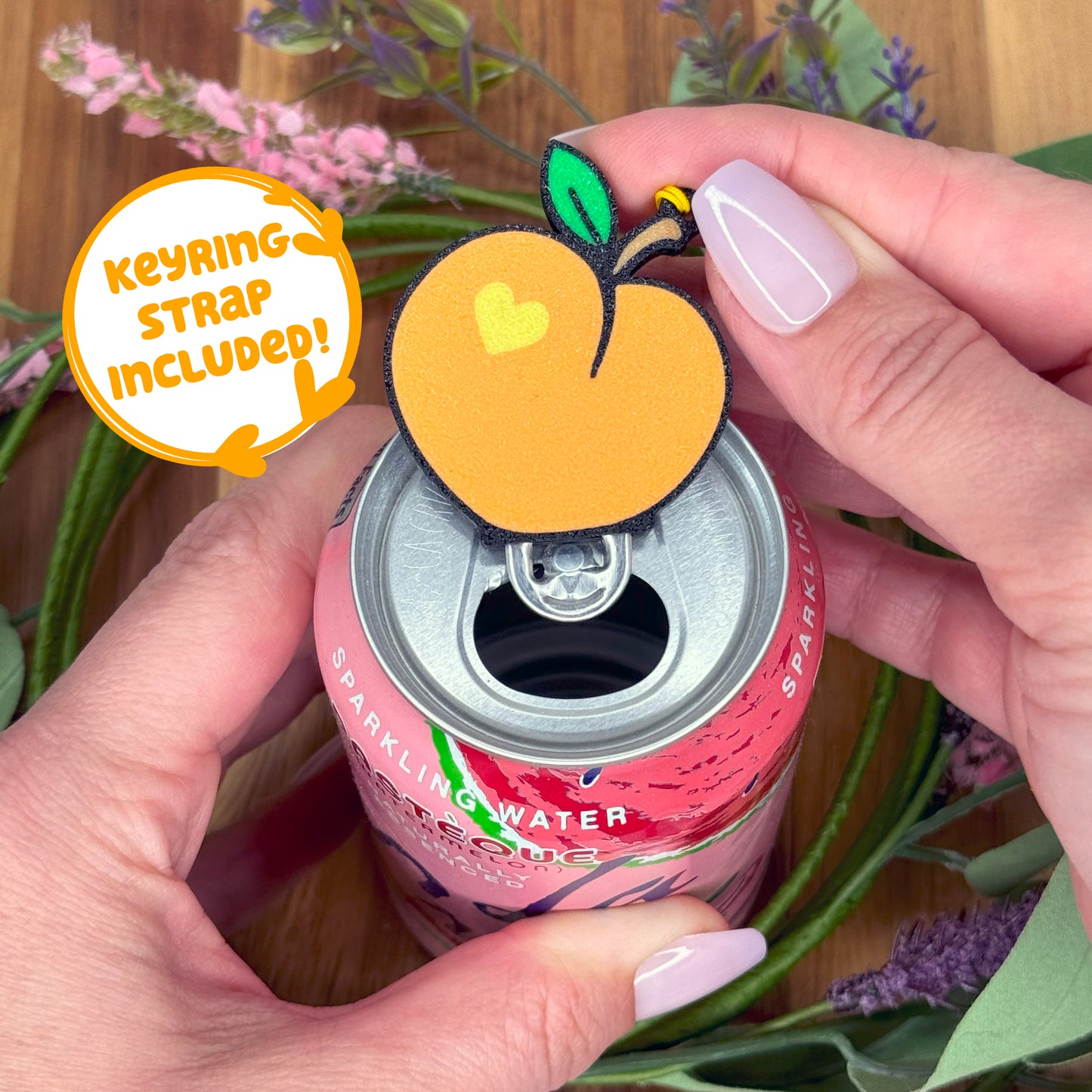 Person holding a can of sparkling water with a peach-shaped Tab Buddy with keyring strap attached, surrounded by flowers.