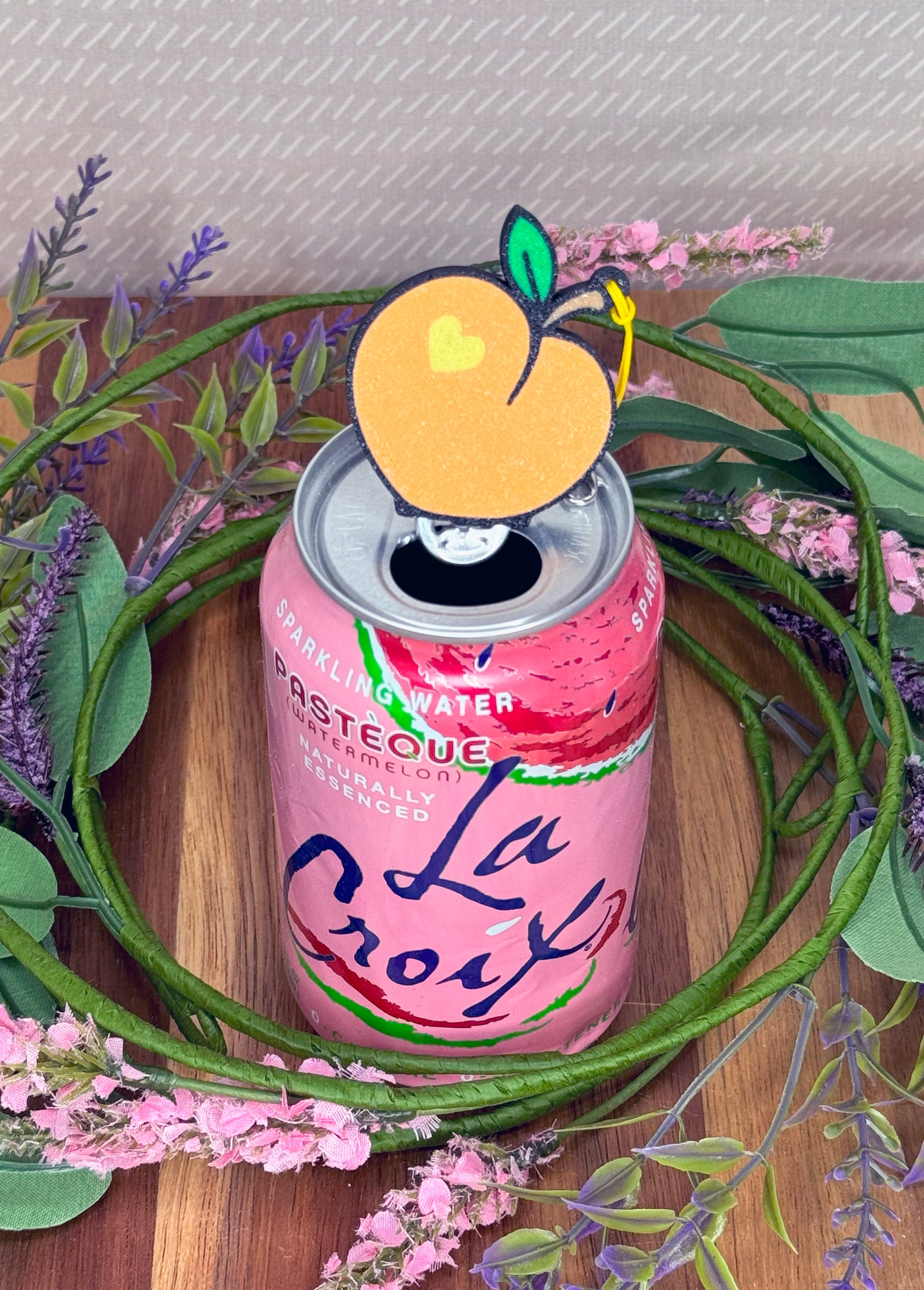 Pink can of sparkling water with a peach-shaped Tab Buddy on top, surrounded by greenery and flowers.