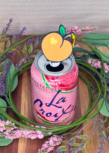 Pink can of sparkling water with a peach-shaped Tab Buddy on top, surrounded by greenery and flowers.