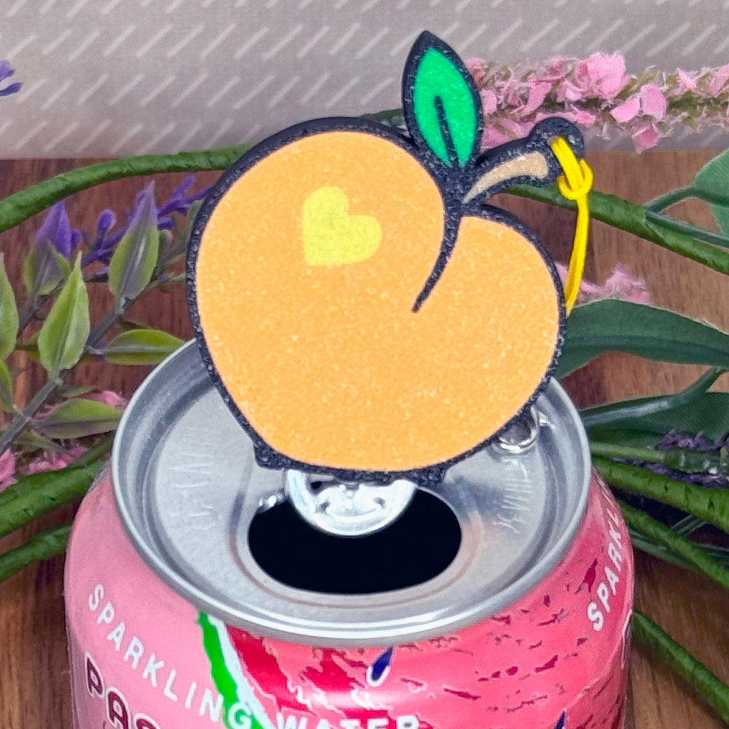 Pink can of sparkling water with a peach Tab Buddy resting on top, surrounded by plants.